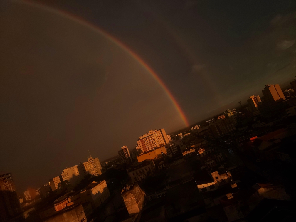 Imagem - Arco-íris vibrante no céu, surgindo de uma cidade com prédios ao fundo, sob um céu parcialmente nublado.